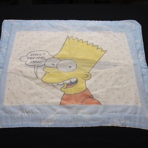 Op de afbeelding: Een witte kussensloop met een blauwe rand heeft een cartoonafbeelding van Bart Simpson uit The Simpsons. De afbeelding toont Bart met een spraakballon die zegt "Sweet Dreams, Man!" De kussensloop heeft een witte achtergrond met kleine rode stippen.