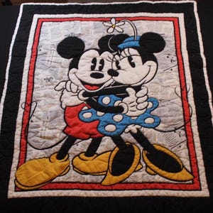 Puede incluir: Una colcha acolchada con Mickey y Minnie Mouse abraz&aacute;ndose. El dise&ntilde;o incluye un borde rojo y blanco, un borde exterior negro y un fondo que se asemeja a una impresi&oacute;n de peri&oacute;dico. Mickey lleva pantalones cortos rojos y zapatos amarillos, mientras que Minnie lleva un vestido azul con lunares blancos.