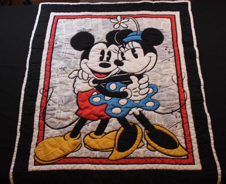 Puede incluir: Una colcha acolchada con Mickey y Minnie Mouse abraz&aacute;ndose. El dise&ntilde;o incluye un borde rojo y blanco, un borde exterior negro y un fondo que se asemeja a una impresi&oacute;n de peri&oacute;dico. Mickey lleva pantalones cortos rojos y zapatos amarillos, mientras que Minnie lleva un vestido azul con lunares blancos.