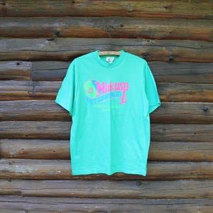 May include: A bright green short-sleeve t-shirt featuring a graphic on the chest with the text Nakusp Hot Springs British Columbia Canada in pink and blue lettering, along with a circular logo design in pink, blue, and green colors.