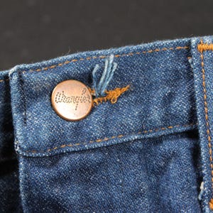 May include: Close-up of a blue denim waistband with a copper-colored button that reads "Wrangler".