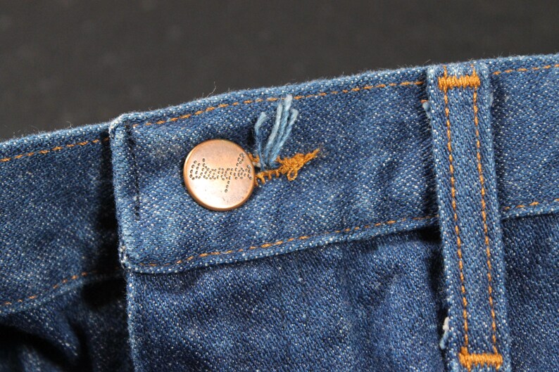 May include: Close-up of a blue denim waistband with a copper-colored button that reads "Wrangler".