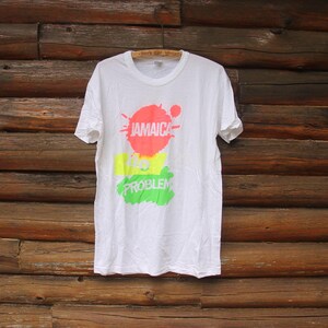 以下が含まれることがあります： 白地にカラフルな3つの円が描かれたグラフィックデザインのTシャツ。円は赤、黄、緑です。円には白い文字で「ジャマイカノー проблем」と書いてあります。