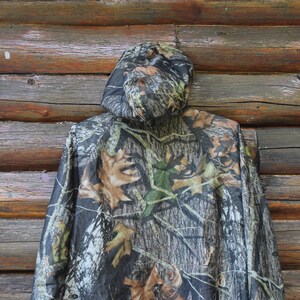 May include: A camouflage hooded jacket with a pattern of brown, green, and black, resembling leaves and branches. The jacket is hanging against a rustic wooden background.