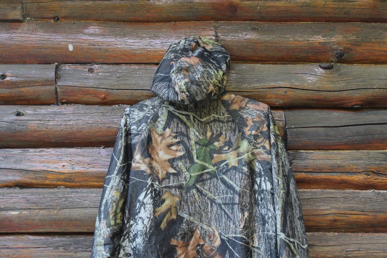 May include: A camouflage hooded jacket with a pattern of brown, green, and black, resembling leaves and branches. The jacket is hanging against a rustic wooden background.