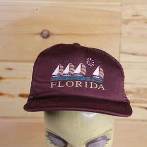 Op de afbeelding: Een donkerrode truckercap met een wit mesh achterpaneel. De voorkant van de cap heeft een afbeelding van vier zeilboten met een zon en sterren in de lucht. De tekst "FLORIDA" is onder de zeilboten gedrukt.