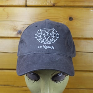May include: A charcoal gray baseball cap featuring a white embroidered logo that reads Vuarnet Sunglasses with a V symbol inside a circle. Below the logo, the text La légende is embroidered in white. The cap has a soft, suede-like texture.