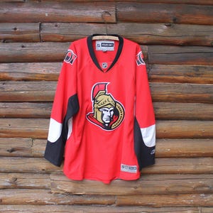 May include: A red hockey jersey with black and white accents. The jersey features a team logo on the front and is hanging on a wooden background. The jersey has long sleeves and a V-neck.
