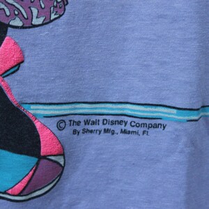 May include: A light purple t-shirt featuring a graphic print with black, teal, and bright pink colors. The text reads: The Walt Disney Company, By Sherry Mfg., Miami, FL. The fabric has a visible weave texture and a horizontal teal line graphic.