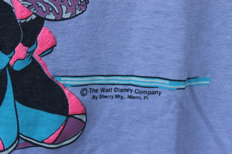 May include: A light purple t-shirt featuring a graphic print with black, teal, and bright pink colors. The text reads: The Walt Disney Company, By Sherry Mfg., Miami, FL. The fabric has a visible weave texture and a horizontal teal line graphic.