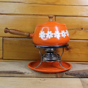 Può includere: Una fonduta arancione con accenti di margherite bianche, un manico e un coperchio in legno. La pentola poggia su un supporto cromato con una base arancione abbinata. Lo sfondo è una parete di legno.