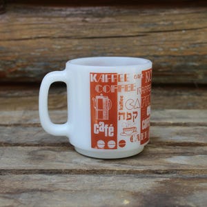 Peut inclure: Mug à café blanc avec une anse, orné de texte et d'illustrations orange liés au café et à la culture des cafés. La tasse comprend les mots "Kaffee", "Coffee" et "Café".