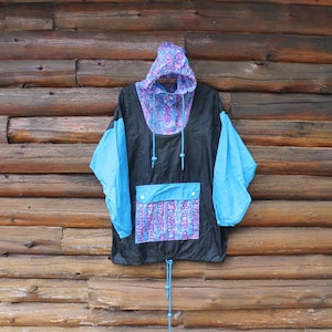 Op de afbeelding: Een vintage zwarte en blauwe anorakjas met capuchon. De jas heeft een patroon in paars en blauw op de capuchon en de zak. De mouwen zijn helderblauw en de jas heeft een trekkoord in de taille.