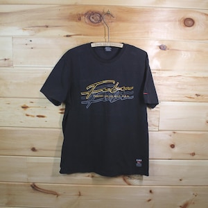May include: Black short-sleeved t-shirt with a crew neck. The shirt features a graphic design in yellow, blue, and white, with the text "FUBU" and "1992 EST. HOLDERS, N.Y.C. USA". The shirt is hanging on a metal hanger.