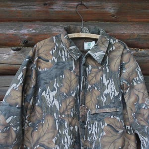 Puede incluir: Chaqueta de camuflaje con cierre de botones, cuello y bolsillo en el pecho. La chaqueta presenta un estampado de formas de hojas marrones, grises y blancas. Cuelga de una percha de madera sobre un fondo de madera.