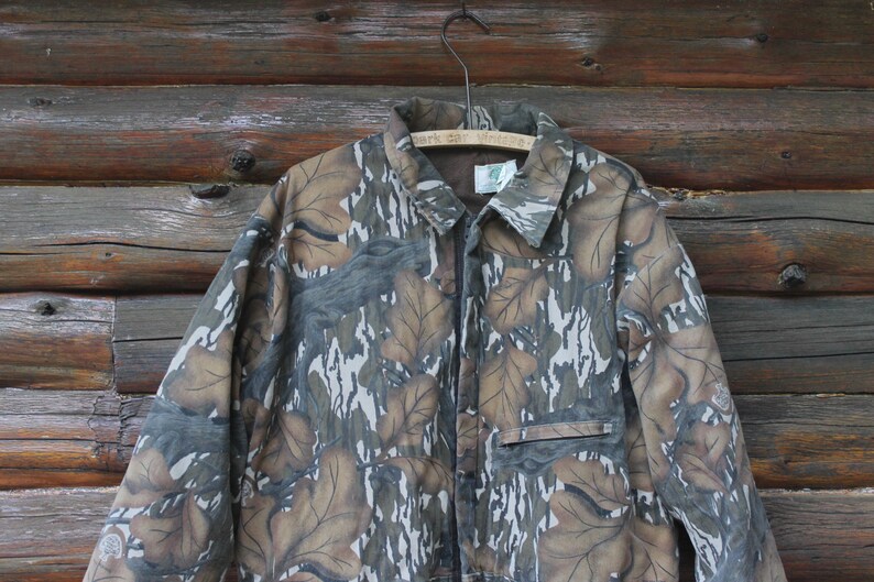 Puede incluir: Chaqueta de camuflaje con cierre de botones, cuello y bolsillo en el pecho. La chaqueta presenta un estampado de formas de hojas marrones, grises y blancas. Cuelga de una percha de madera sobre un fondo de madera.