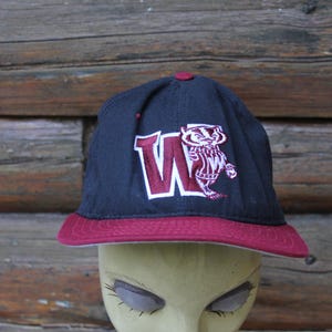 May include: Navy blue baseball cap with a maroon brim and button. The cap features a maroon "W" with a badger mascot logo. The hat is displayed on a mannequin head against a wooden background.