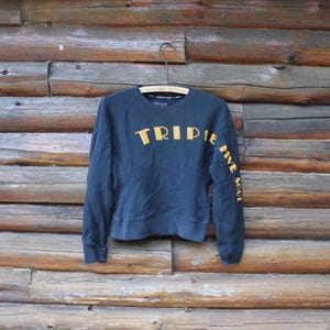 May include: A navy blue sweatshirt with the word "TRIPLE" in yellow block letters across the chest and "FIVE SOUL" down the left sleeve. The sweatshirt is hanging on a wooden hanger against a rustic wooden background.