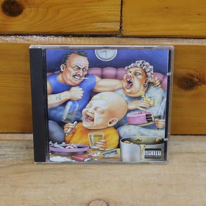 May include: A compact disc in a clear plastic jewel case featuring a colorful cartoon illustration of a family scene. The artwork depicts a man in a blue tank top, a woman, and a baby, with various items including a cake, drinks, and a pot.
