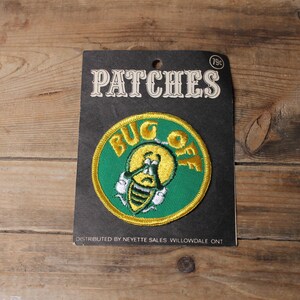 May include: A vintage patch on a black backing, with the words "BUG OFF" around a cartoon insect. The patch is green and yellow with white accents. The backing reads "PATCHES" in a vintage font.