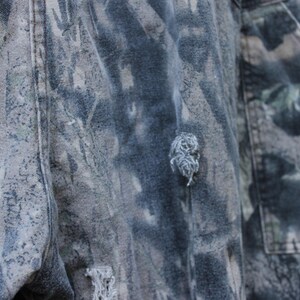 May include: Close-up of camouflage trousers with a torn area. The trousers feature a pattern of greens, browns, and greys. The fabric appears worn, with visible fraying around the tear, suggesting a rugged aesthetic.