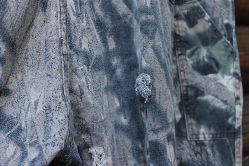 May include: Close-up of camouflage trousers with a torn area. The trousers feature a pattern of greens, browns, and greys. The fabric appears worn, with visible fraying around the tear, suggesting a rugged aesthetic.