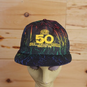 May include: Black baseball cap with a rainbow and black print. The cap has a yellow embroidered logo that says "DuPont Canada 50 Years 100% Nylon 1992 Kingston Site".