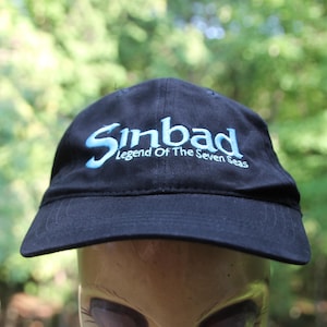 Peut inclure: Casquette de baseball noire avec l'inscription "Sinbad Legend Of The Seven Seas" en bleu clair. La casquette a une visière incurvée et est faite d'un tissu foncé. L'arrière-plan est une vue floue de feuillage vert.