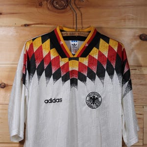 May include: White soccer jersey with a black, red, and yellow geometric pattern on the shoulders. The jersey has the Adidas logo and the German national team crest.