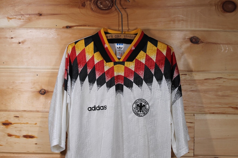 May include: White soccer jersey with a black, red, and yellow geometric pattern on the shoulders. The jersey has the Adidas logo and the German national team crest.