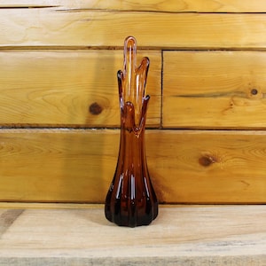 Peut inclure: Un vase en verre ambré, de forme élancée, avec une base évasée et un corps cannelé. Le vase a une forme organique unique avec trois extensions pointues vers le haut. Le vase est posé sur un fond en bois.