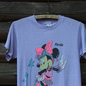 May include: A light purple short-sleeve t-shirt featuring a graphic of Minnie Mouse wearing pink sunglasses and a bow. The character is accessorized with a beaded necklace and bracelets. The text Florida is printed in black near the character.