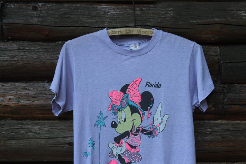 May include: A light purple short-sleeve t-shirt featuring a graphic of Minnie Mouse wearing pink sunglasses and a bow. The character is accessorized with a beaded necklace and bracelets. The text Florida is printed in black near the character.
