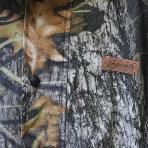 May include: Close-up of a camouflage pattern fabric with a brown leather Coleman label. The fabric features a mix of green, brown, and gray tones, with a button visible on the left side. The overall theme is outdoor apparel.