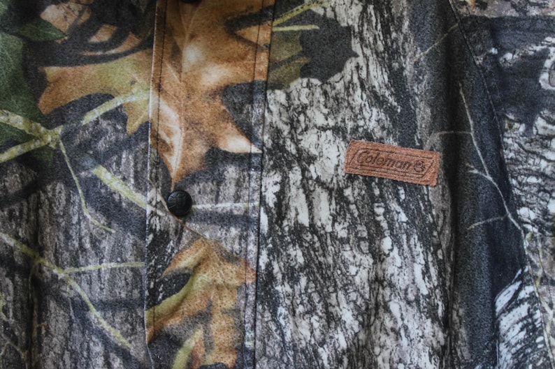 May include: Close-up of a camouflage pattern fabric with a brown leather Coleman label. The fabric features a mix of green, brown, and gray tones, with a button visible on the left side. The overall theme is outdoor apparel.