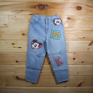 Peut inclure: Jean clair avec des écussons Mickey Mouse. Les écussons comprennent un Mickey en forme de cœur, la lettre "M" dans un carré à pois et la lettre "K" dans un carré rose. Le jean est suspendu à un cintre en métal.