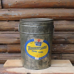 May include: A vintage metal pail with a lid and handle. The pail has a faded, aged appearance and features a large oval label with the text "Tenderflake Pure Lard" in blue and yellow. The background is a rustic wooden wall.