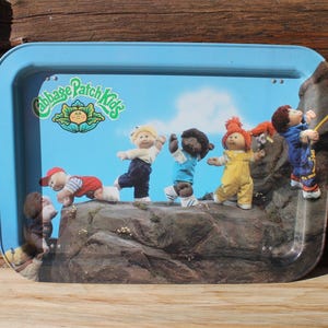 May include: A rectangular, light blue metal tray with a Cabbage Patch Kids design. The tray shows several Cabbage Patch Kids dolls climbing a rocky surface against a blue sky background. The Cabbage Patch Kids logo is in the upper left corner.