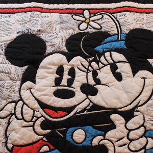 Puede incluir: Manta acolchada con Mickey y Minnie Mouse abraz&aacute;ndose. Los personajes en blanco y negro se encuentran sobre un fondo de peri&oacute;dico. La manta tiene un borde rojo y un reverso negro.
