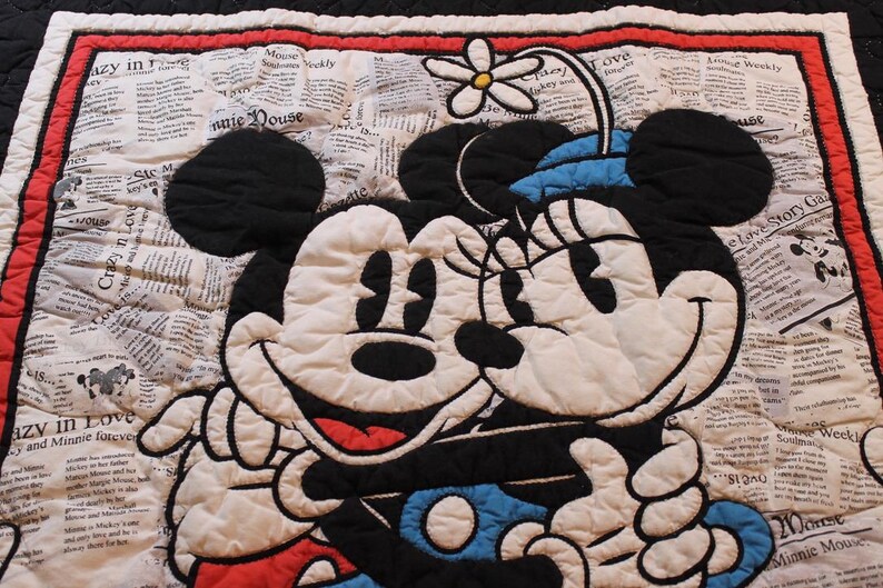 Puede incluir: Manta acolchada con Mickey y Minnie Mouse abraz&aacute;ndose. Los personajes en blanco y negro se encuentran sobre un fondo de peri&oacute;dico. La manta tiene un borde rojo y un reverso negro.