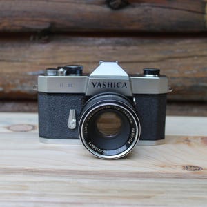 Op de afbeelding: Een vintage Yashica FX-3 35mm filmcamera met een zwarte behuizing en zilveren accenten. De lens is gemarkeerd met "Auto Yashinon-DX 1:2 f=50mm". De camera staat op een licht houten oppervlak tegen een achtergrond van houten planken.