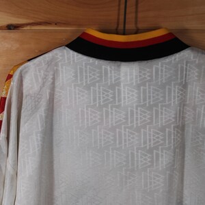 May include: White long-sleeve shirt with a black, red, and yellow collar. The shirt has a repeating pattern of white triangles on the front.