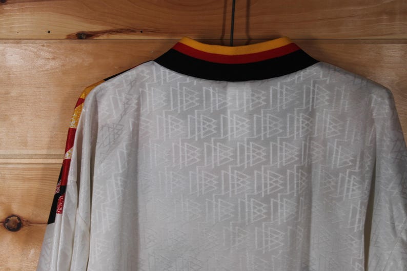 May include: White long-sleeve shirt with a black, red, and yellow collar. The shirt has a repeating pattern of white triangles on the front.