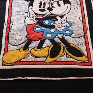 Puede incluir: Manta acolchada con Mickey y Minnie Mouse abraz&aacute;ndose. Los personajes est&aacute;n sobre un fondo de peri&oacute;dico. La manta tiene un borde rojo, un ribete negro y un borde blanco. Mickey lleva pantalones cortos rojos y zapatos amarillos, mientras que Minnie lleva un vestido azul.