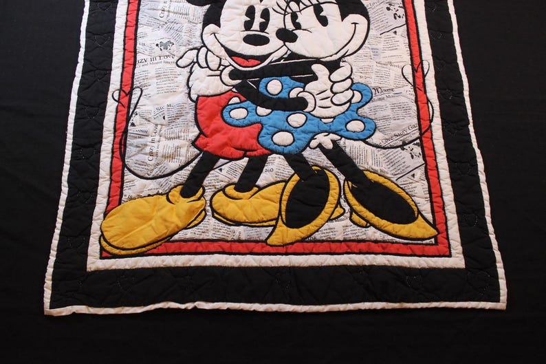 Puede incluir: Manta acolchada con Mickey y Minnie Mouse abraz&aacute;ndose. Los personajes est&aacute;n sobre un fondo de peri&oacute;dico. La manta tiene un borde rojo, un ribete negro y un borde blanco. Mickey lleva pantalones cortos rojos y zapatos amarillos, mientras que Minnie lleva un vestido azul.