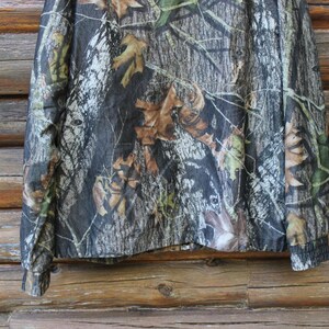 May include: Camouflage long-sleeve shirt with a woodland pattern. The shirt features a mix of brown, green, and gray tones, mimicking a forest environment. The sleeves have ribbed cuffs. The shirt is displayed against a wooden background.