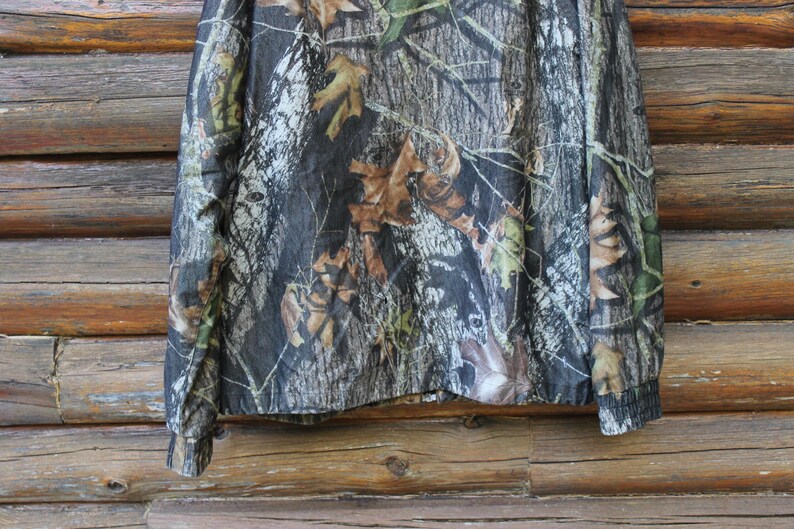 May include: Camouflage long-sleeve shirt with a woodland pattern. The shirt features a mix of brown, green, and gray tones, mimicking a forest environment. The sleeves have ribbed cuffs. The shirt is displayed against a wooden background.