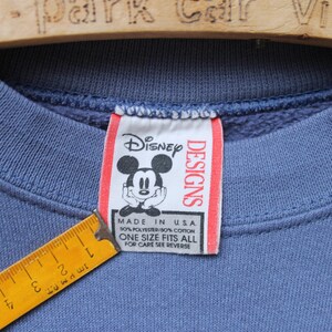 Puede incluir: Primer plano de un cuello de sudadera azul con una etiqueta de Disney Designs con Mickey Mouse. La etiqueta dice "Made in USA" y "Talla &uacute;nica". Una regla amarilla es parcialmente visible.