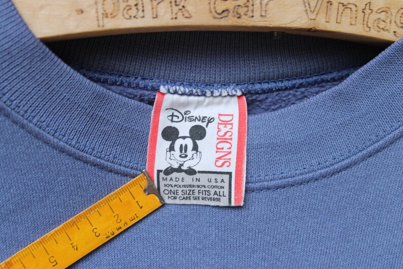Puede incluir: Primer plano de un cuello de sudadera azul con una etiqueta de Disney Designs con Mickey Mouse. La etiqueta dice "Made in USA" y "Talla &uacute;nica". Una regla amarilla es parcialmente visible.