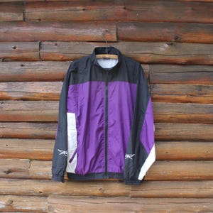 Könnte beinhalten: Vintage schwarze, lila und weiße Sportjacke mit durchgehendem Reißverschluss. Die Jacke hat ein Colorblock-Design mit weißen Akzenten an den Ärmeln und einem kleinen Logo. Die Jacke hängt an einem Holzkleiderbügel vor einem Holzgrund.